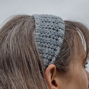 May include: A gray knitted headband with a textured pattern. The headband is worn on a person's head.