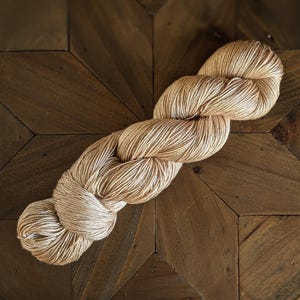 May include: A skein of beige yarn with a subtle shimmer. The yarn is wound into a loose coil and is lying on a wooden surface.