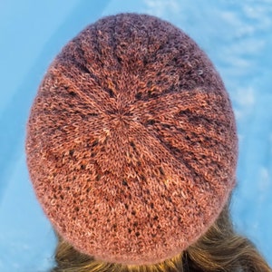 May include: A close-up of a knitted beanie in a deep mauve color. The beanie has a textured stitch pattern and is made of a soft, warm yarn.