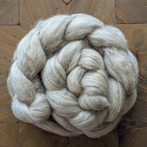 May include: A large, braided bundle of light gray wool roving. The wool is tightly spun and has a soft, textured appearance. The roving is arranged in a circular pattern, with the braid visible throughout. The background is a wooden surface.