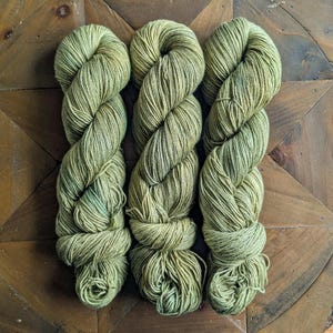 May include: Three skeins of green yarn are displayed, each twisted into a rope-like shape. The yarn has a variegated color, with shades of olive and hints of blue. The yarn is arranged on a dark wooden surface with a geometric pattern.