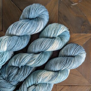 May include: Close-up of several skeins of yarn in shades of blue, teal, and white. The yarn is twisted and arranged on a dark wooden surface, showcasing the texture and color variations. Ideal for knitting and crochet projects.