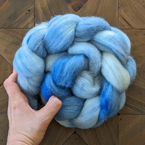 May include: A hand-spun ball of blue and white wool yarn. The yarn is arranged in a braided pattern, creating a textured and visually appealing look.