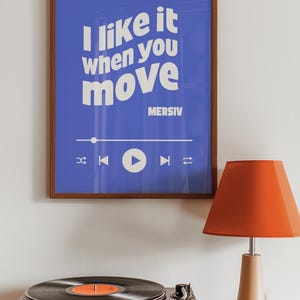Peut inclure: Une affiche encadr&eacute;e avec un fond bleu et du texte blanc qui dit "J'aime &ccedil;a quand tu bouges" avec le nom de l'artiste "MERSIV" sous le texte. L'affiche a un bouton de lecture et d'autres commandes de lecteur de musique en bas. Un tourne-disque vintage avec un disque noir sur la platine est au premier plan. Une lampe rouge avec une base beige est sur le c&ocirc;t&eacute; droit de l'image.
