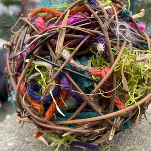 May include: A round, woven sphere made of twigs and colorful yarn. The yarn is in shades of pink, orange, green, blue, and purple. The sphere is sitting on a gray, textured surface.