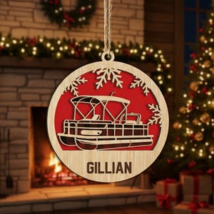 May include: Circular wooden Christmas ornament with a pontoon boat design and the name "GILLIAN". The boat is set against a red backdrop, framed by snowflakes. The ornament is suspended by twine. The background includes a fireplace and Christmas tree.