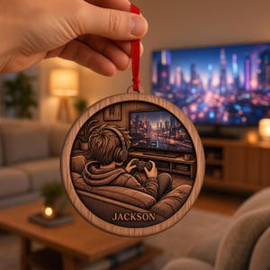 May include: A round wooden ornament featuring a laser-engraved illustration of a person wearing headphones and playing a video game on a television. The name JACKSON is engraved at the bottom. The ornament hangs from a thin red ribbon.