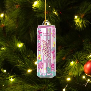Puede incluir: Adorno navide&ntilde;o a rayas rosas y blancas con forma de lata de bebida energ&eacute;tica. La lata presenta el texto "Cotton Candy" y "Alani Energy" con ilustraciones florales y de p&aacute;jaros. La parte inferior de la lata dice "KIRA 2025".