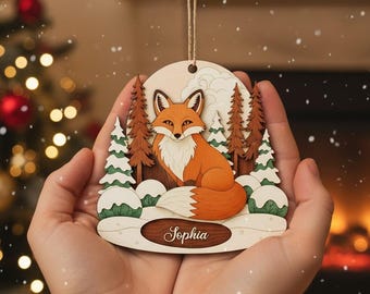 May include: Wooden Christmas ornament featuring a fox in a snowy forest scene. The fox is orange and white, with a detailed design. The ornament has a light brown background with the name "Sophia" engraved on a wooden plaque. A string is attached for hanging.