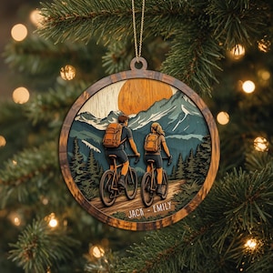 May include: A round wooden ornament featuring a layered illustration of two people riding bicycles on a dirt path towards mountains under a large orange sun. The names JACK and EMILY are printed in white at the bottom of the circular wooden frame.