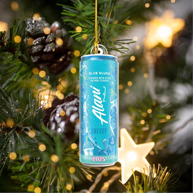 Puede incluir: Una lata azul de bebida energ&eacute;tica Alani Nu, etiquetada como "Blue Slush", cuelga de un hilo dorado en un &aacute;rbol de Navidad. La lata presenta un dise&ntilde;o azul claro con el nombre de la marca y la palabra "Energy". El fondo incluye agujas de pino, pi&ntilde;as y luces navide&ntilde;as.