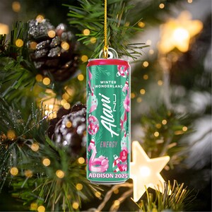 Puede incluir: Adorno navide&ntilde;o con forma de lata de bebida energ&eacute;tica Alani Nu. La lata es verde y roja y presenta las palabras "Winter Wonderland" y "Energy". Elementos decorativos incluyen flamencos y adornos. Cuelga de un hilo dorado.