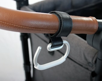 May include: A silver stroller hook attached to a brown leather handlebar with a black leather strap.