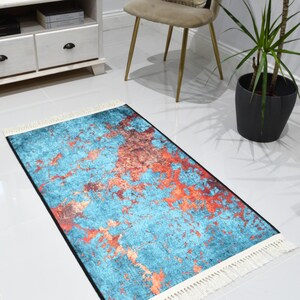 May include: A rectangular rug with a teal and orange abstract pattern. The rug has a black border and white fringe on the short ends. The rug is placed on a white tiled floor.