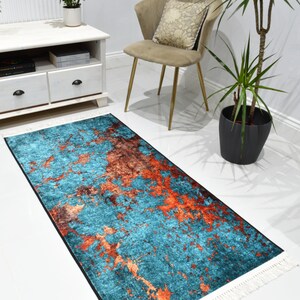 May include: A rectangular rug with a teal and orange abstract design. The rug has a black border and white fringe on the short ends. The rug is placed on a white floor.