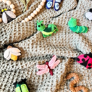 May include: A collection of felt insect and butterfly decorations in various colors, including green, blue, pink, red, and brown. The decorations are arranged on a textured, light brown fabric background.