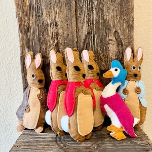 May include: A collection of felt stuffed animals, including several brown rabbits with pink-lined ears and red scarves, and a white and pink duck with a blue hat. The toys are displayed on a rustic wooden shelf.
