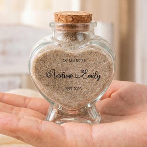May include: A heart-shaped glass bottle with a cork stopper, filled with light brown sand. The front of the glass is printed with the text 28 MARCH, the names Andrew & Emily, and EST. 2023 in a black serif and script font combination.