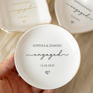 May include: Three white ceramic ring dishes with gold trim. One is square, one heart-shaped, and one round. Each dish has the word "engaged" and a date, with names. A hand holds the round dish. A diamond ring is visible.