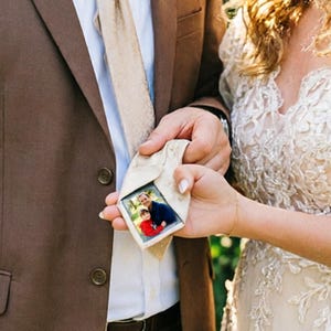 Könnte beinhalten: Eine beige Krawatte mit einem kleinen gerahmten Foto eines Mannes und eines Kindes. Die Krawatte wird von zwei Personen gehalten, eine in einem braunen Anzug und die andere in einem weißen Spitzenkleid. Das Foto ist in einem kleinen, quadratischen Rahmen.