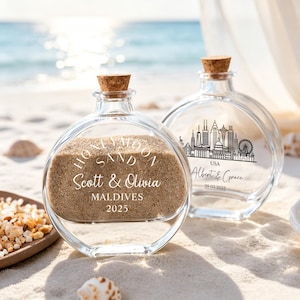 May include: Two round clear glass bottles with cork stoppers filled with sand. The front bottle features white text reading HONEYMOON SAND, Scott & Olivia, MALDIVES, 2025. The back bottle displays a black city skyline, USA, Albert & Grace, 09.03.2025.