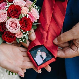 Puede incluir: Una corbata roja con un patrón de rayas diagonales, con una fotografía enmarcada. La corbata se sostiene cerca de un ramo de rosas rojas y rosas. La fotografía muestra a dos personas. La corbata está siendo sostenida por dos personas.