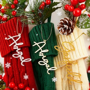 May include: Three knit Christmas stockings in red, green, and cream, each featuring a hanging name ornament. The red and green stockings display the name Abigail in white and gold script, while the cream stocking displays the name Garfield in gold glitter script.