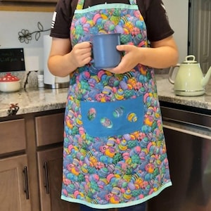 May include: A colorful apron with a blue pocket and a vibrant pattern of Easter egg-like designs in shades of blue, pink, yellow, and green. The apron is trimmed with a light blue border and features a matching blue pocket with decorative cutouts.