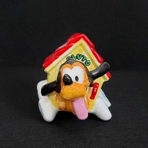 May include: A ceramic figurine of Pluto the dog from Disney, wearing a red and yellow doghouse on his head. Pluto is depicted with a brown body, black ears, and a pink tongue sticking out.
