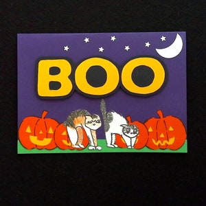 May include: A Halloween-themed card with the word "BOO" in yellow letters with black outlines. The background is purple with white stars and a crescent moon. Two cartoon cats and three orange jack-o'-lanterns are at the bottom.