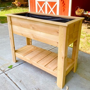 May include: A wooden raised garden bed with a black liner and a lower shelf. The planter is made of natural wood and has a rustic look.