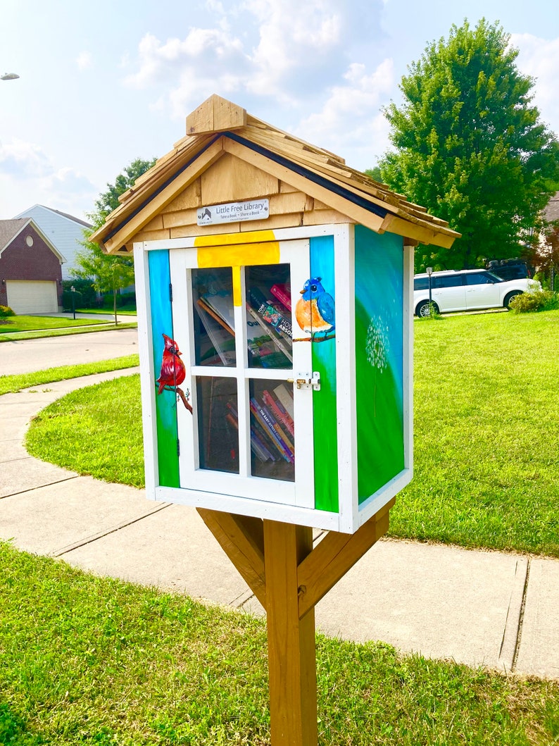 Puede incluir: Una peque&ntilde;a biblioteca gratuita de madera con un exterior pintado de azul, verde y blanco. La biblioteca tiene una peque&ntilde;a ventana con estantes llenos de libros. El techo es marr&oacute;n y tiene un dise&ntilde;o de tejas. La biblioteca est&aacute; pintada con un p&aacute;jaro azul, un cardenal y un diente de le&oacute;n.