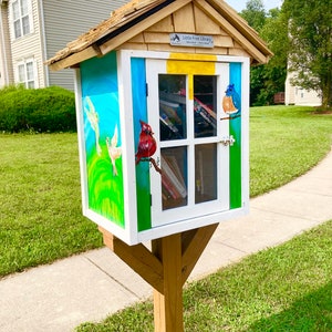 Puede incluir: Una peque&ntilde;a biblioteca gratuita de madera pintada con un cielo azul, hierba verde y p&aacute;jaros coloridos. La biblioteca tiene una puerta blanca y una ventana con libros en su interior. El letrero en la parte delantera dice "Little Free Library - Toma un libro, comparte un libro".