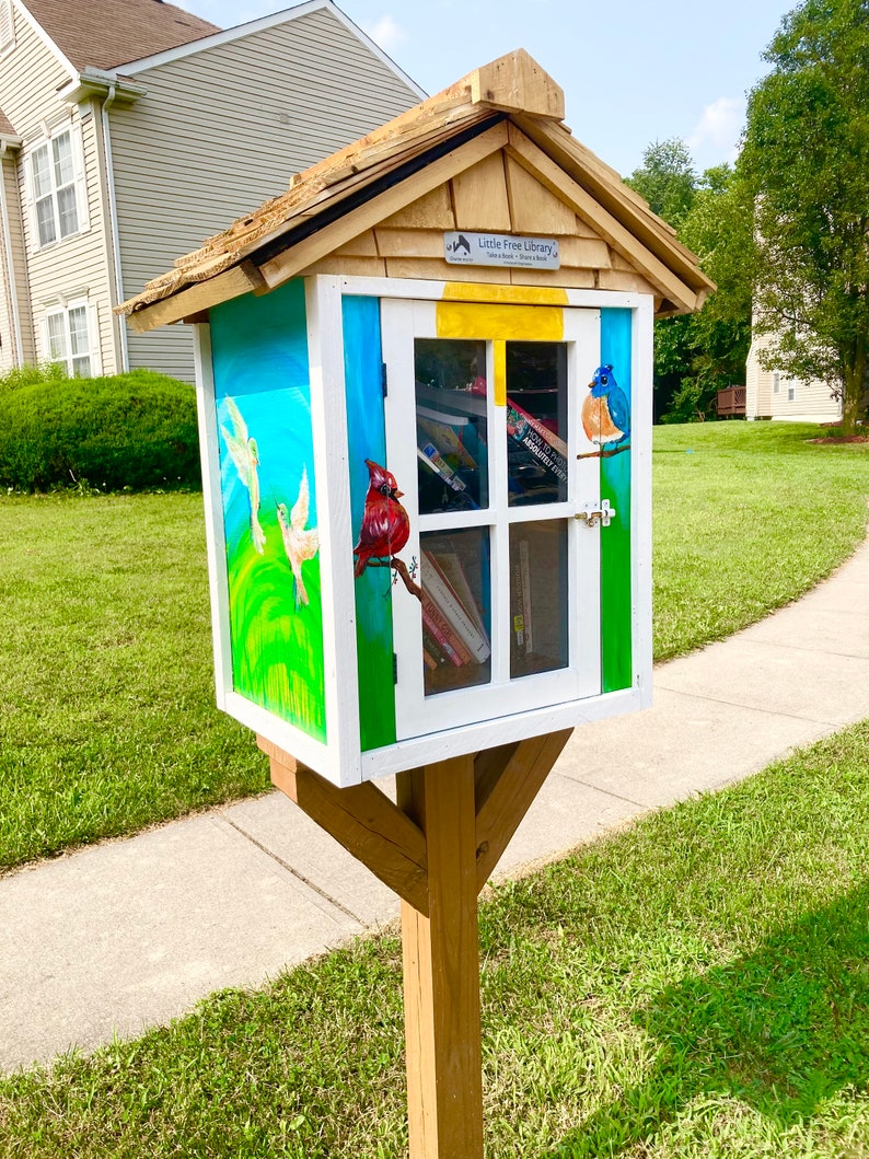 Puede incluir: Una peque&ntilde;a biblioteca gratuita de madera pintada con un cielo azul, hierba verde y p&aacute;jaros coloridos. La biblioteca tiene una puerta blanca y una ventana con libros en su interior. El letrero en la parte delantera dice "Little Free Library - Toma un libro, comparte un libro".