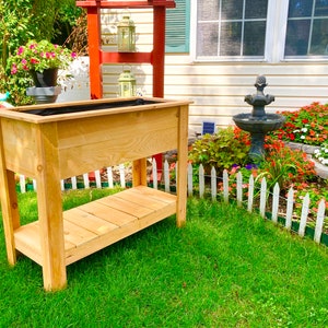 May include: A wooden planter box with a black liner, sitting on a wooden stand with a lower shelf. The planter is in a garden setting with a white picket fence, green grass, and flowers.