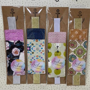 May include: Four fabric bookmarks with different patterns, each attached to a cardboard backing with an anchor design. The patterns include floral, apple, and geometric designs. Each bookmark has a label that says "Anchor Handmade Goods."