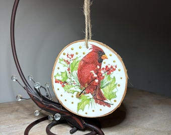 May include: A round wooden ornament featuring a red cardinal bird perched on a branch with red berries and green leaves. The ornament has a natural wood grain edge and is decorated with gold dots. It hangs from a jute twine string.