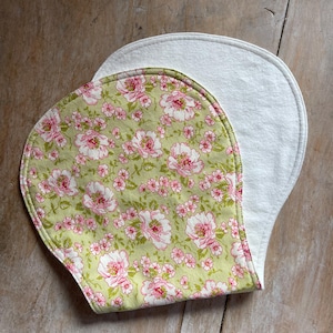 May include: Two baby bibs. One bib features a floral pattern with pink and white flowers on a light green background. The other bib is solid white. Both bibs have a rounded shape and are made of soft fabric.