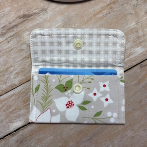 May include: Open fabric card holder with a floral design. The holder features a light gray background with white flowers, red accents, and green leaves. The interior is light gray with a small checkered pattern. A blue card is visible inside.