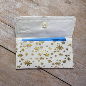 May include: A white fabric card holder with a gold snowflake pattern. The interior is white, and a blue card is visible inside. The holder has a snap closure.