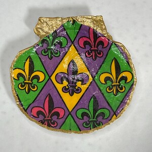 May include: A seashell painted with a purple, green, and yellow diamond pattern and fleur-de-lis designs. The shell is trimmed with gold paint.