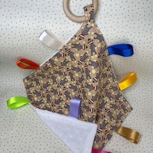 May include: A beige baby sensory toy with a wooden ring and multiple colorful satin ribbons. The toy is decorated with a pattern of monkeys wearing red hats.