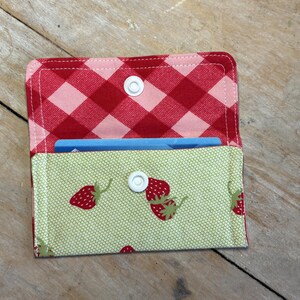 May include: Open fabric card holder with a red and pink gingham pattern on the inside and a light green strawberry print on the outside. The card holder is secured with a white snap closure and holds a blue card.
