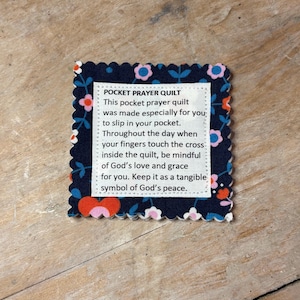 May include: A navy blue pocket prayer quilt with a scalloped edge, adorned with a floral pattern in pink, red, and white. The quilt features a white rectangular label with text describing its purpose and meaning. The text reads "POCKET PRAYER QUILT".
