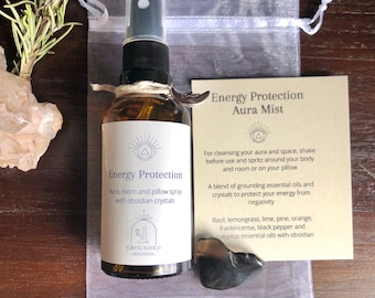 Può includere: Una bottiglia di vetro trasparente con un'etichetta marrone che recita "Energy Protection Aura, room and pillow spray with obsidian crystals". La bottiglia è in una borsa di organza bianca con un sasso di ossidiana nera. L'etichetta include anche il testo "GROUNDED Wellness".