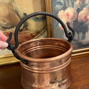 Peut inclure: Un chaudron rond en cuivre avec une finition martelée polie et une large ouverture. Le récipient est doté d'une anse épaisse et incurvée en fer sombre, fixée sur les côtés par des supports métalliques décoratifs en forme de crochet.