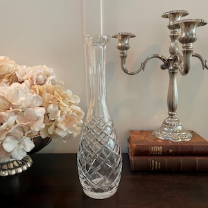 May include: A clear, diamond-cut glass vase with a slender neck and a bulbous base. The vase is displayed on a dark wooden surface, next to a silver candelabra and a stack of antique books. The vase is empty.