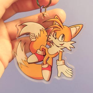 Könnte beinhalten: Ein klarer Acryl-Schlüsselanhänger mit der Figur Tails aus der Sonic the Hedgehog-Serie. Der Schlüsselanhänger hat ein farbenfrohes Design mit orangefarbenen, weißen und roten Akzenten. Er ist an einem Metallverschluss befestigt.