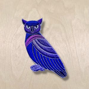 Pode incluir: Remendo de coruja bordada em tons de azul e roxo. A coruja apresenta costuras detalhadas, com uma faixa roxa no peito e costuras brancas que delineiam o desenho. Em um fundo de grão de madeira clara.