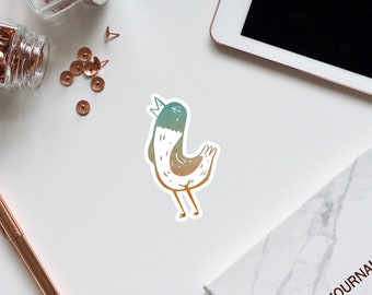May include: A sticker featuring a whimsical bird illustration. The bird has a white body with gradient colors of teal, brown, and orange. It is wearing a small crown. The sticker is on a white surface with office supplies.